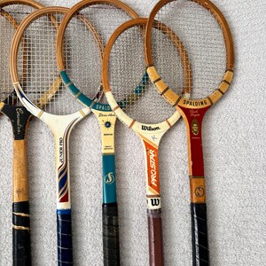 May include: A collection of vintage wooden tennis rackets. The rackets have wooden frames, string netting, and various grip colors. Some rackets have brand names like Spalding and Wilson. The grips are wrapped in black, brown, and blue tape.