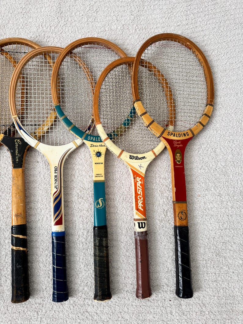 May include: A collection of vintage wooden tennis rackets. The rackets have wooden frames, string netting, and various grip colors. Some rackets have brand names like Spalding and Wilson. The grips are wrapped in black, brown, and blue tape.