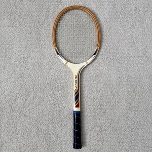 May include: Vintage tennis racket with a wooden frame and a white handle. The racket features a cream-colored head with a brown border and the text "JUNIOR PRO" on the handle. The grip is wrapped in dark blue tape.