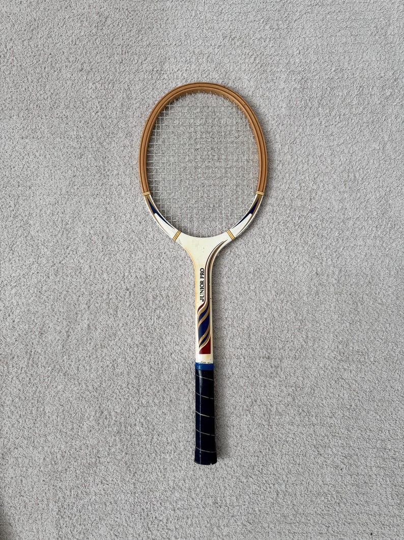 May include: Vintage tennis racket with a wooden frame and a white handle. The racket features a cream-colored head with a brown border and the text "JUNIOR PRO" on the handle. The grip is wrapped in dark blue tape.