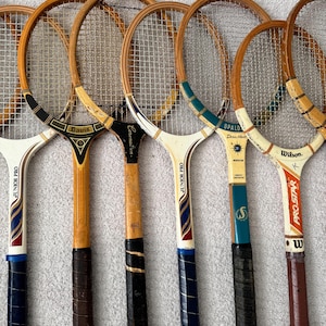 May include: A collection of vintage tennis rackets with wooden frames and stringed faces. The rackets feature various handle designs and brand names, including "Davis," "Spalding," and "Wilson." The grips are wrapped in leather or tape, and the rackets show signs of wear.
