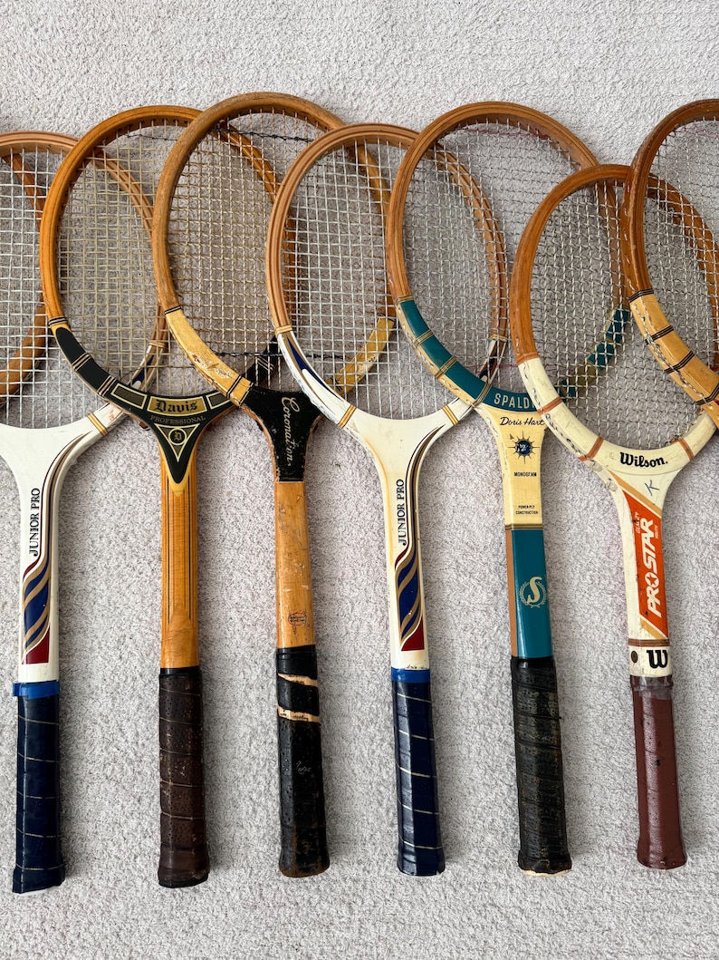 May include: A collection of vintage tennis rackets with wooden frames and stringed faces. The rackets feature various handle designs and brand names, including "Davis," "Spalding," and "Wilson." The grips are wrapped in leather or tape, and the rackets show signs of wear.