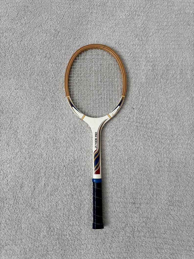 May include: Vintage tennis racket with a wooden frame and a white handle. The racket has the text "JUNIOR PRO" printed on the handle. The grip is black with a blue band. The racket's frame is brown and the strings are silver.