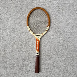 May include: Vintage Wilson Pro Star tennis racket with a wooden frame and a brown grip. The racket has a white and orange handle with the Wilson logo and Pro Star text. The racket is in good condition.