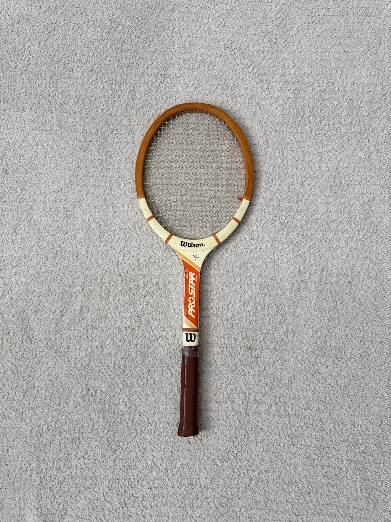 May include: Vintage Wilson Pro Star tennis racket with a wooden frame and a brown grip. The racket has a white and orange handle with the Wilson logo and Pro Star text. The racket is in good condition.