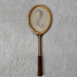 May include: Vintage wooden tennis racket with a brown leather grip. The racket head is oval-shaped with a stringed net. The frame is light brown with black and white stripes. The text "LA VITESSE" is printed on the side of the racket.