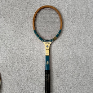 May include: Vintage Spalding tennis racket with a wooden frame and a teal and gold handle. The racket has a black grip and the text "Spalding Doris Hart" is visible on the head.