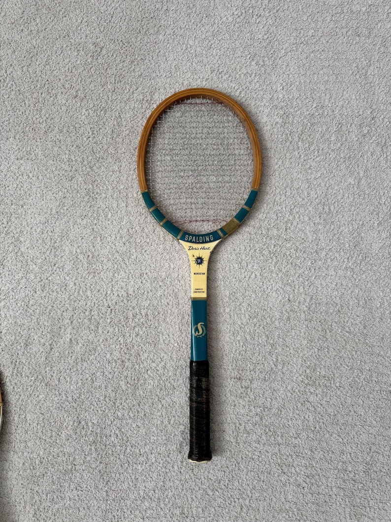 May include: Vintage Spalding tennis racket with a wooden frame and a teal and gold handle. The racket has a black grip and the text "Spalding Doris Hart" is visible on the head.