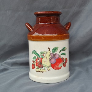 May include: A brown and white ceramic milk jug with a fruit design. The jug has a brown lid and two handles. The fruit design includes a pear, grapes, and strawberries.