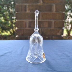 May include: A clear crystal bell with a faceted design and a long, slender handle. The bell has a gold sticker on the side.