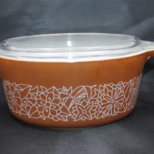May include: A brown ceramic casserole dish with a clear glass lid. The dish has a white floral design around the rim.