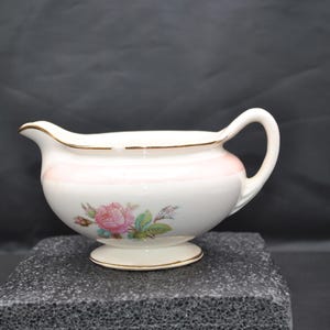 May include: White ceramic gravy boat with a pink floral design and gold trim. The gravy boat has a handle and a spout for easy pouring. The floral design features pink roses and green leaves. The gravy boat sits on a small pedestal base.