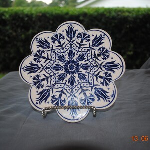 May include: A scalloped, circular ceramic plate featuring a white base with a detailed, symmetrical blue floral pattern of tulips and leaves arranged in a mandala-like design. The plate is supported by a silver-toned metal wire display stand.