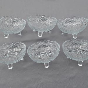 May include: Six clear glass footed bowls with intricate etched designs. The bowls are all the same size and shape, and they are arranged in a row on a gray surface.