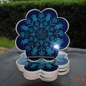 May include: A stack of five flower-shaped ceramic coasters featuring a deep blue background with a repeating turquoise floral pattern. The top coaster is displayed upright on a silver wire stand, showcasing the intricate symmetrical design.