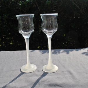 May include: Two clear, tulip-shaped wine glasses with long, slender stems and frosted white bases. The glasses are made of clear glass and are displayed on a light gray surface, with a dark green background.