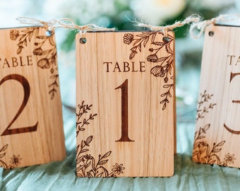 May include: Wooden table number signs with floral designs. Each sign displays a number from 1 to 3, with the word "TABLE" above. The signs are made of light-colored wood and are decorated with engraved floral patterns.