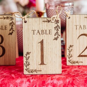 Puede incluir: Tres letreros de madera con números de mesa con diseños florales. Los letreros están etiquetados como "Mesa 1", "Mesa 2" y "Mesa 3".