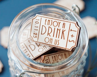 May include: A glass jar filled with wooden drink tickets that read "Enjoy a Drink on Us" and the date "10.11.23". The jar sits on a blue surface, with scattered tickets and white flowers. The tickets are light brown.