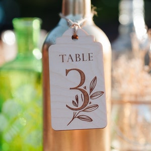 Può includere: Una bottiglia di vino dipinta d'oro con un'etichetta di legno che recita "TABLE 3" con un design botanico. La bottiglia fa parte di un'impostazione del tavolo per matrimoni o eventi. Una bottiglia verde è sullo sfondo.