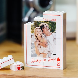 May include: A white building block structure shaped like a playing card, featuring a printed photograph of a couple, the date 12.11.2026, and the red text Lucky in Love. Two smaller matching block pieces with red heart symbols sit in front.