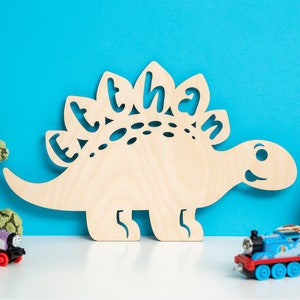 May include: A wooden dinosaur cutout with the name "Ethan" cut out of the back. The dinosaur is light brown and is sitting on a white surface in front of a blue wall. There are three toy trains in the foreground, one purple, one blue, and one green.