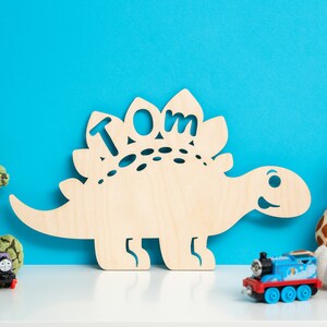 May include: A wooden dinosaur cutout with the name "TOM" cut out of the back. The dinosaur is light brown and has a friendly face. The dinosaur is sitting on a white shelf in front of a blue wall. There are toy trains and stuffed animals on the shelf.