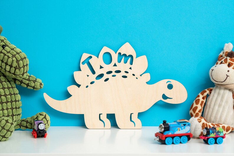 May include: A wooden dinosaur cutout with the name "TOM" cut out of the back. The dinosaur is light brown and has a friendly face. The dinosaur is sitting on a white shelf in front of a blue wall. There are toy trains and stuffed animals on the shelf.
