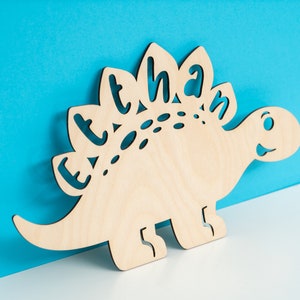 May include: A wooden cutout of a dinosaur with the name "Ethan" spelled out on its back. The dinosaur is light brown and has a friendly face.