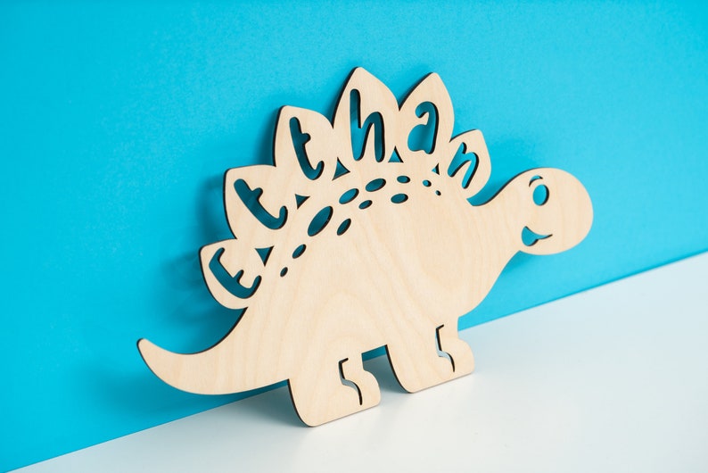 May include: A wooden cutout of a dinosaur with the name "Ethan" spelled out on its back. The dinosaur is light brown and has a friendly face.
