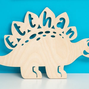 May include: A wooden cutout of a dinosaur with the name "Ethan" spelled out on its back. The dinosaur is light brown and has a friendly face.