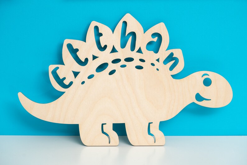 May include: A wooden cutout of a dinosaur with the name "Ethan" spelled out on its back. The dinosaur is light brown and has a friendly face.
