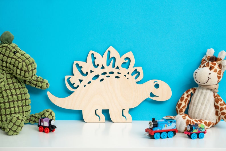 May include: A wooden dinosaur cutout with the name "Ethan" cut out of the back. The dinosaur is light brown and is sitting on a white surface in front of a blue wall. There are three toy trains in the foreground, one purple, one blue, and one green.