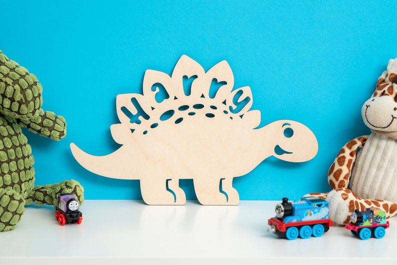 May include: A wooden dinosaur cutout with the name "Harry" cut out of the back. The dinosaur is light brown and has a friendly face. The dinosaur is sitting on a white surface with a green plush dinosaur, a blue toy train, and a giraffe plush toy.