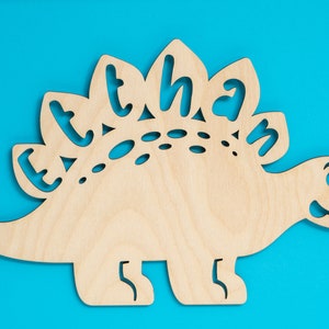 May include: A wooden cutout of a dinosaur with the name "Ethan" spelled out on its back. The dinosaur is light brown and has a blue background.