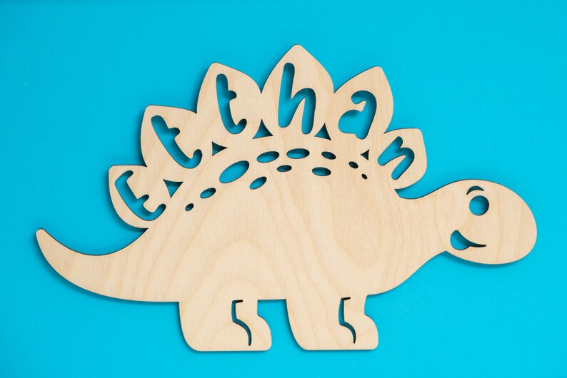 May include: A wooden cutout of a dinosaur with the name "Ethan" spelled out on its back. The dinosaur is light brown and has a blue background.