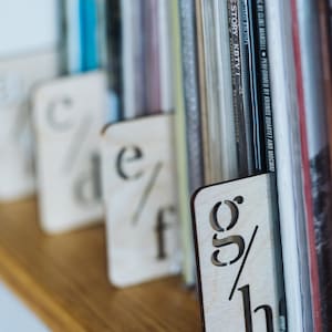 Puede incluir: Separadores de libros de madera con las letras 'c', 'd', 'e', 'f', 'g' y 'h' talladas en ellos. Los separadores de libros est&aacute;n colocados entre filas de libros en un estante de madera.