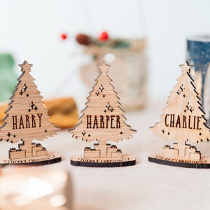 May include: Three wooden Christmas tree place cards with names "Harry", "Harper", and "Charlie" engraved. Each tree has a star and decorative cutouts. The base of each tree reads "Merry Christmas".