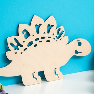May include: A wooden dinosaur cutout with the name "Harry" cut out of the back. The dinosaur is light brown and has a friendly face.