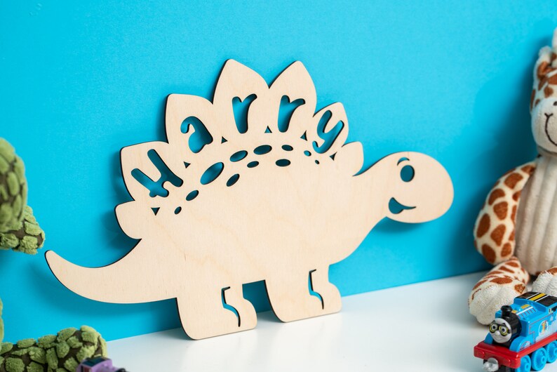 May include: A wooden dinosaur cutout with the name "Harry" cut out of the back. The dinosaur is light brown and has a friendly face.