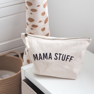 May include: A white canvas zippered pouch with the text "MAMA STUFF" printed in black. The pouch is sitting on a white surface.