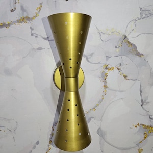 Pode incluir: Uma arandela de parede dourada com duas cúpulas em forma de cone. As cúpulas têm um padrão perfurado e estão inclinadas em direções opostas.
