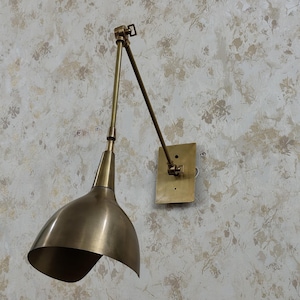 May include: A wall-mounted lamp with a brass finish, featuring a rectangular backplate, an adjustable articulated arm with two segments, and a wide, bell-shaped shade. The metal surface has a brushed texture, and the design includes visible hinges.