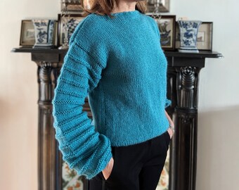 May include: A teal blue knitted sweater with a relaxed fit and long sleeves. The sleeves have a unique textured design.