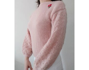 May include: A pale pink knitted sweater with a delicate floral embroidery on the chest. The sweater has a round neckline and long sleeves with a textured pattern.