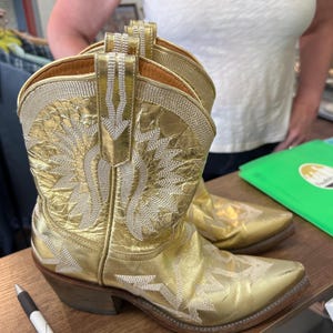 May include: A pair of metallic gold cowboy boots featuring intricate white stitched patterns on the shaft and toe. The boots have a pointed toe, a stacked wooden heel, and a pull tab at the top of the shaft, showcasing a shiny finish.