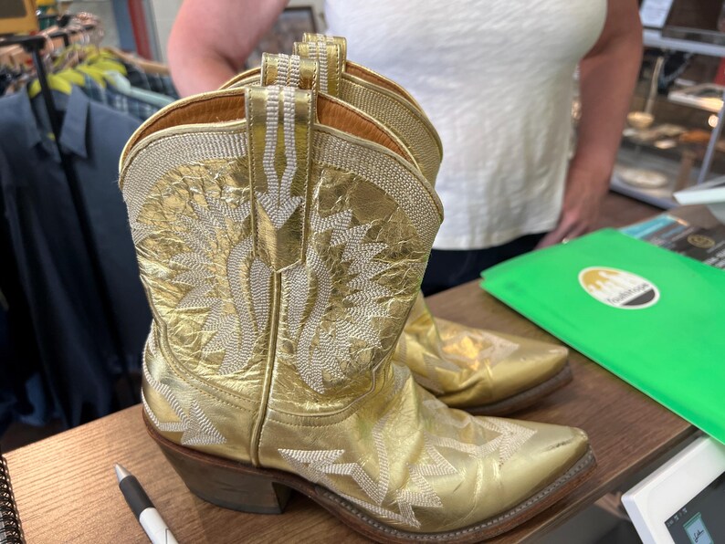 May include: A pair of metallic gold cowboy boots featuring intricate white stitched patterns on the shaft and toe. The boots have a pointed toe, a stacked wooden heel, and a pull tab at the top of the shaft, showcasing a shiny finish.