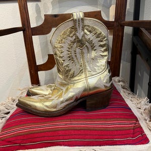 May include: A metallic gold leather cowboy boot featuring intricate white stitched patterns across the shaft and foot. The boot has a pointed toe, a stacked wooden heel, and a pull tab at the top of the shaft, showcasing detailed embroidery.