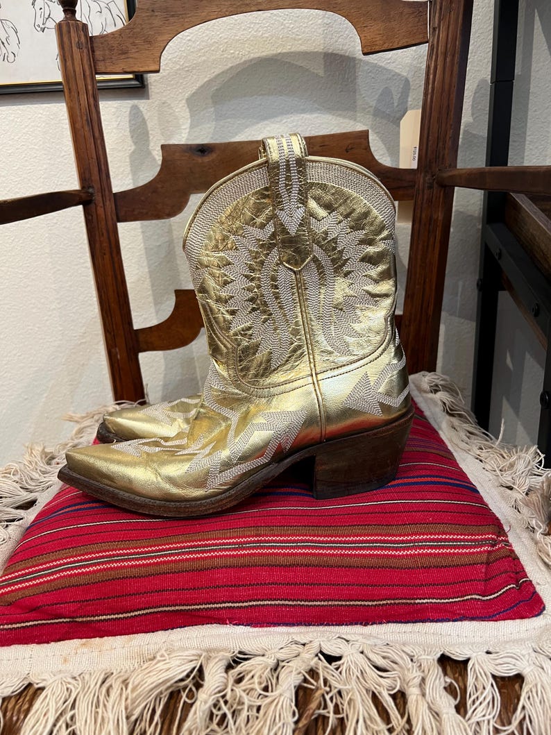 May include: A metallic gold leather cowboy boot featuring intricate white stitched patterns across the shaft and foot. The boot has a pointed toe, a stacked wooden heel, and a pull tab at the top of the shaft, showcasing detailed embroidery.