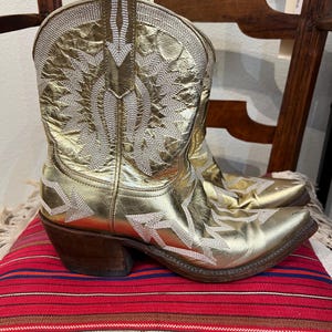 May include: A metallic gold cowboy boot featuring a pointed toe, a stacked wooden heel, and intricate white stitched patterns across the shaft and vamp. The boot has a textured finish and a pull tab at the top of the shaft for easy wear.